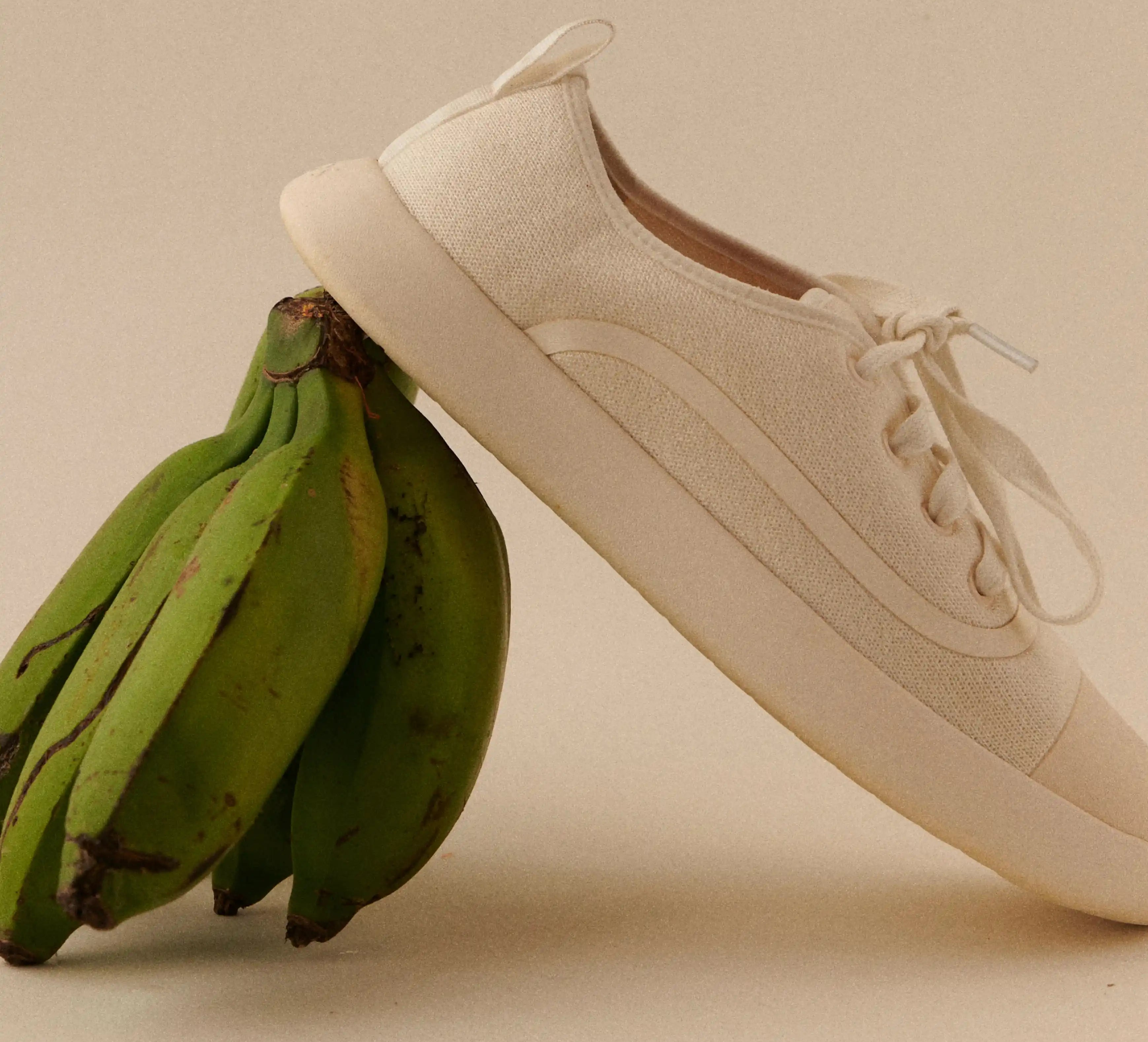 White Juntos sneaker leaning against green bananas on beige background.