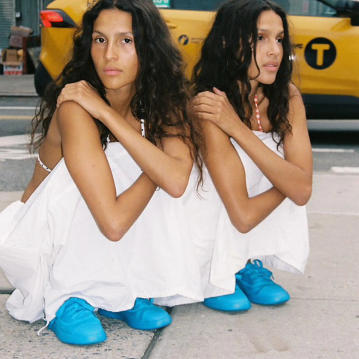 Twin sisters in white dresses wearing Juntos blue sneakers sitting on city street with yellow taxi.